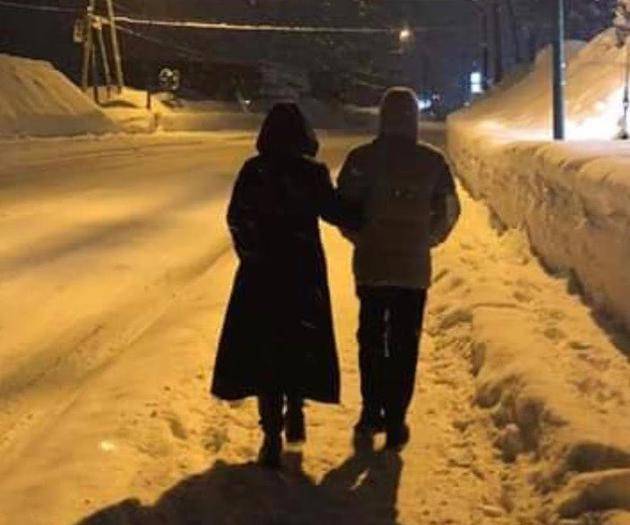 两人在晚上的雪地上行走，两人挽着胳膊，路灯下的身影显得格外温馨与浪漫。