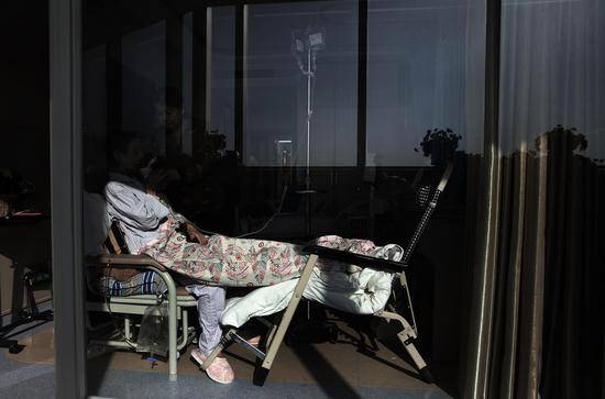 2018年11月12日，北京大学首钢医院安宁疗护中心，一间病房里，一位女士一边输液一边透过玻璃晒着太阳。护工正在边上拿着杯子，让她通过吸管来喝水。新京报记者尹亚飞摄