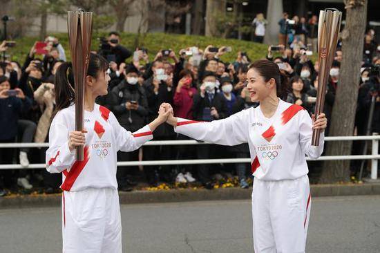 2020年2月15日，东京奥运会圣火传递大使石原里美（右）在彩排中与临时担任“火炬手”的工作人员握手相互鼓励。当日，东京奥组委在日本东京都羽村市举行奥运会火炬接力彩排。照片中，远处的观众大多数都戴起了口罩。图/新华