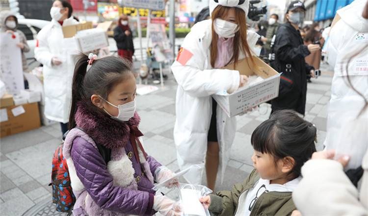 ▲2月29日，在日本东京池袋站前，一名华侨华人小志愿者（左）向当地小朋友发放儿童用口罩｜新华社发