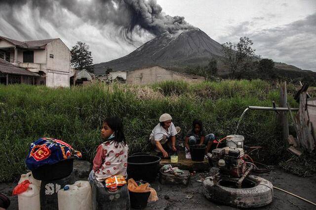 ↑2020年8月14日，当地居民在印度尼西亚北苏门答腊省处于喷发状态的锡纳朋火山脚下洗衣服。新华社发（达马尼克摄）