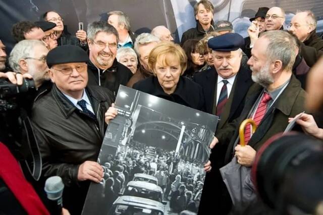 ▲当地时间2009年11月9日，德国柏林，原苏联最高领导人米哈伊尔·戈尔巴乔夫(左)、德国总理默克尔(中)和前波兰总统莱赫·瓦文萨在柏林纪念柏林墙倒塌20周年的仪式上，在他们通过萨姆桥时，手持一幅人们越过布斯布鲁克边境桥的签名版画。图/视觉中国
