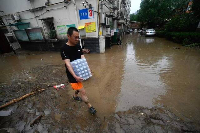 7月31日，北京门头沟，一男子抱着一箱饮用水走在积水的街道上。