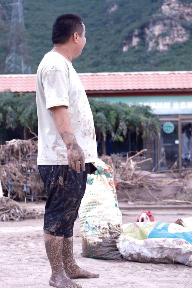 8月4日，洪水退去，淤泥还在，一位小伙子赤脚站在路边，身旁是刚刚收拾好的衣服和杂物。新京报记者曹晶瑞摄