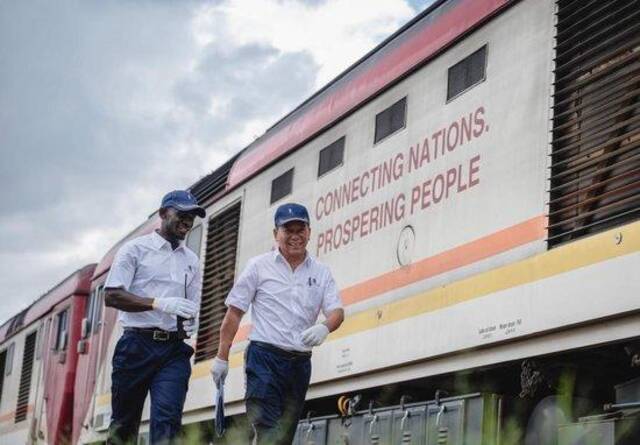 2023年5月23日，在肯尼亚内罗毕，非洲之星铁路运营公司机辆部的火车驾驶员蒋立平（右）与学员霍勒斯从一辆印有“连接国家走向繁荣”标语的蒙内铁路机车旁走过。新华社记者王冠森摄